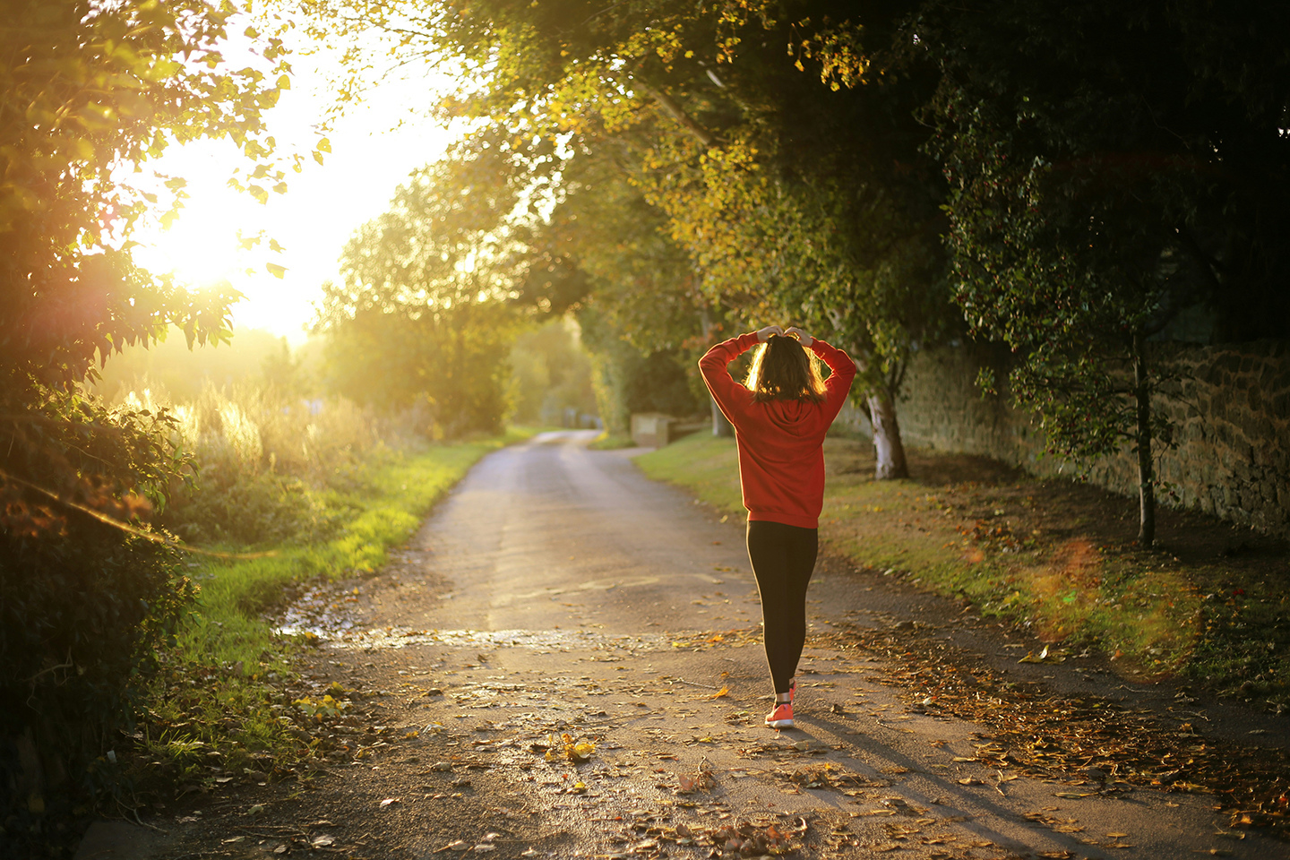 A woman in a red sweater walks down a sunlit path surrounded by trees, holding her hair up as the golden light filters through the leaves.