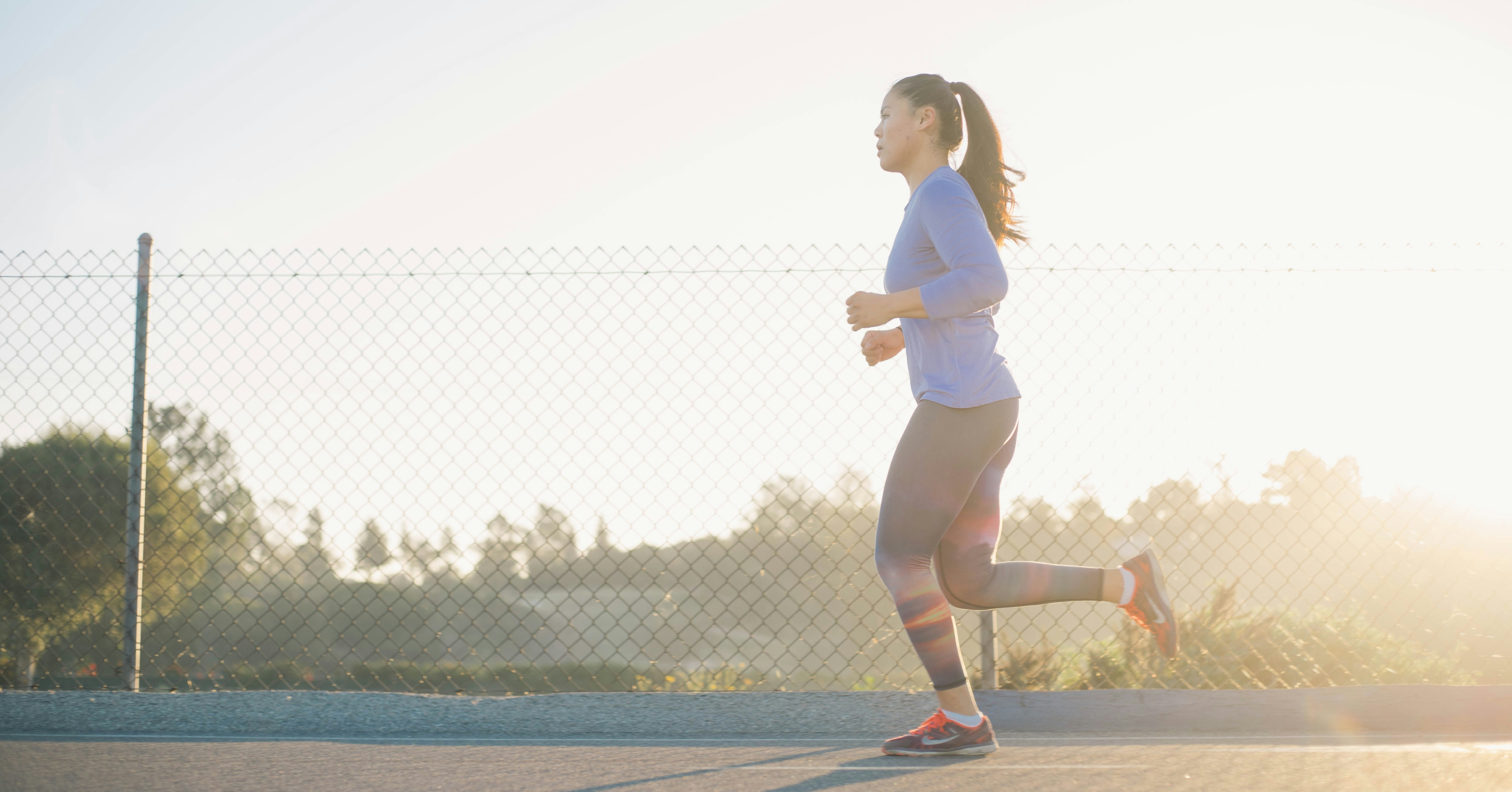 A woman in athletic wear runs outdoors along a path near a chain-link fence, with the sun shining brightly in the background