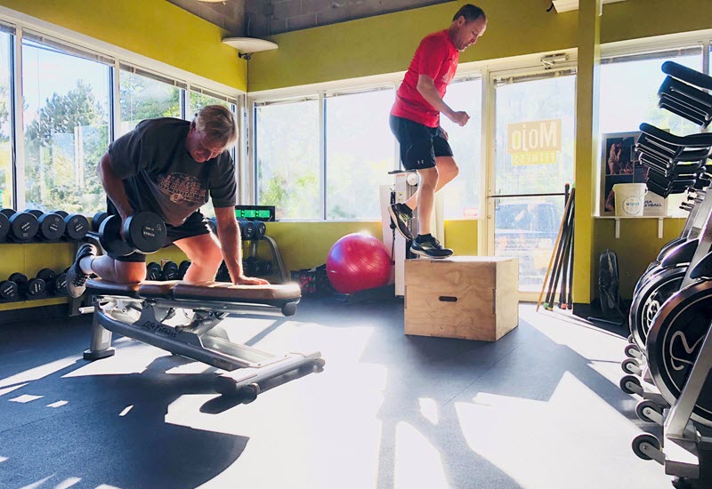 Two men work out in a brightly lit gym, one doing dumbbell exercises on a bench while the other performs box jumps, showcasing strength and cardio training.