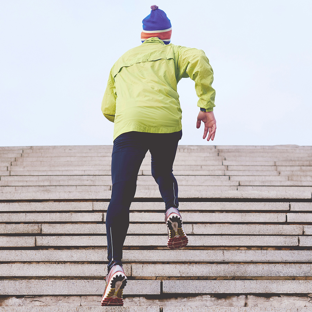 A person , jogging up a flight of concrete steps. They are dressed in a bright yellow jacket, dark athletic pants, and colorful running shoes. A striped beanie cap tops their winter jogging outfit, suggesting a cold-weather exercise scene. The focus is on active lifestyle and physical fitness, captured in an urban setting.