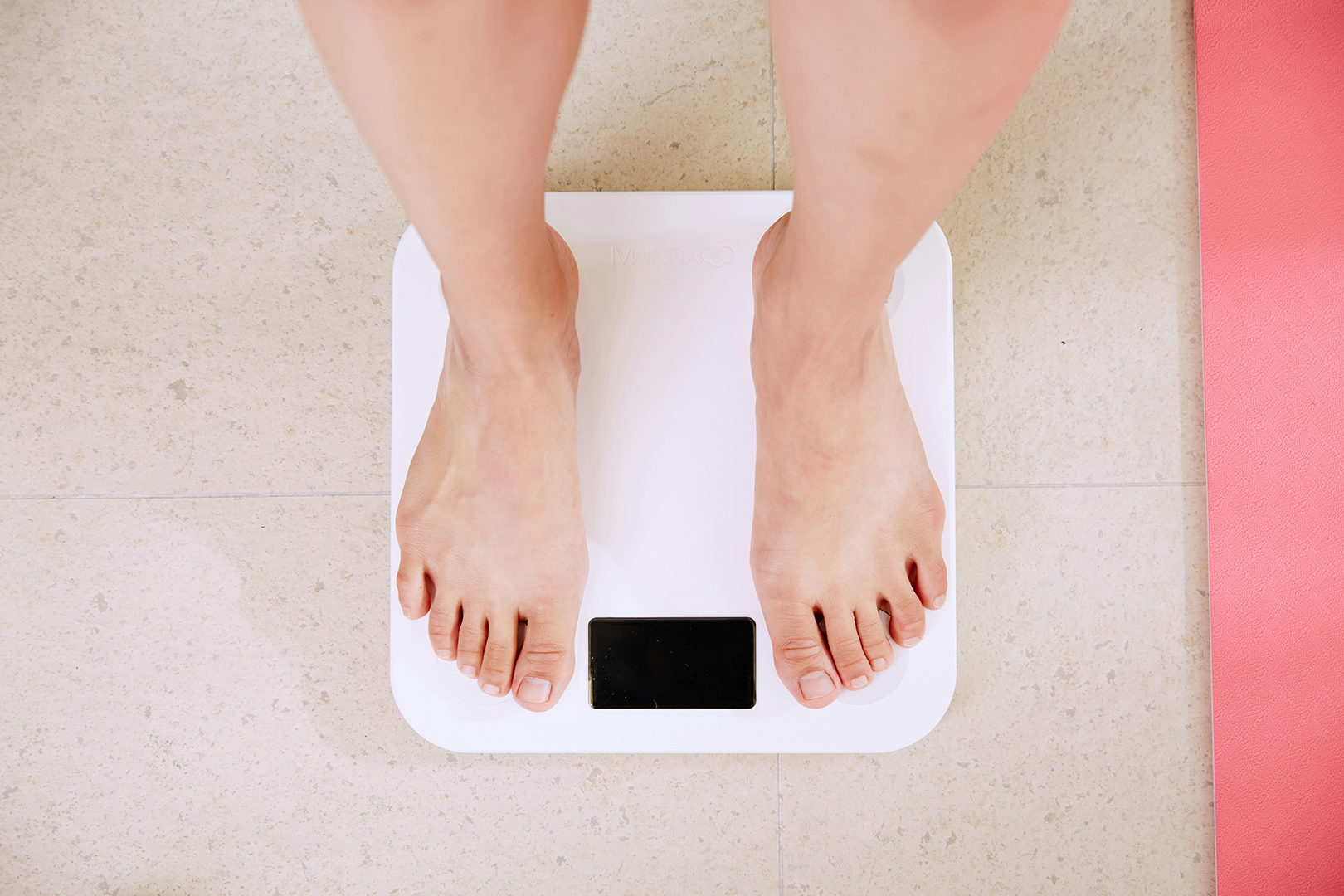 A person's bare feet standing on a white digital scale, with a pink yoga mat visible beside the scale on a textured tile floor. The scale display is not visible, indicating a focus on weight measurement.