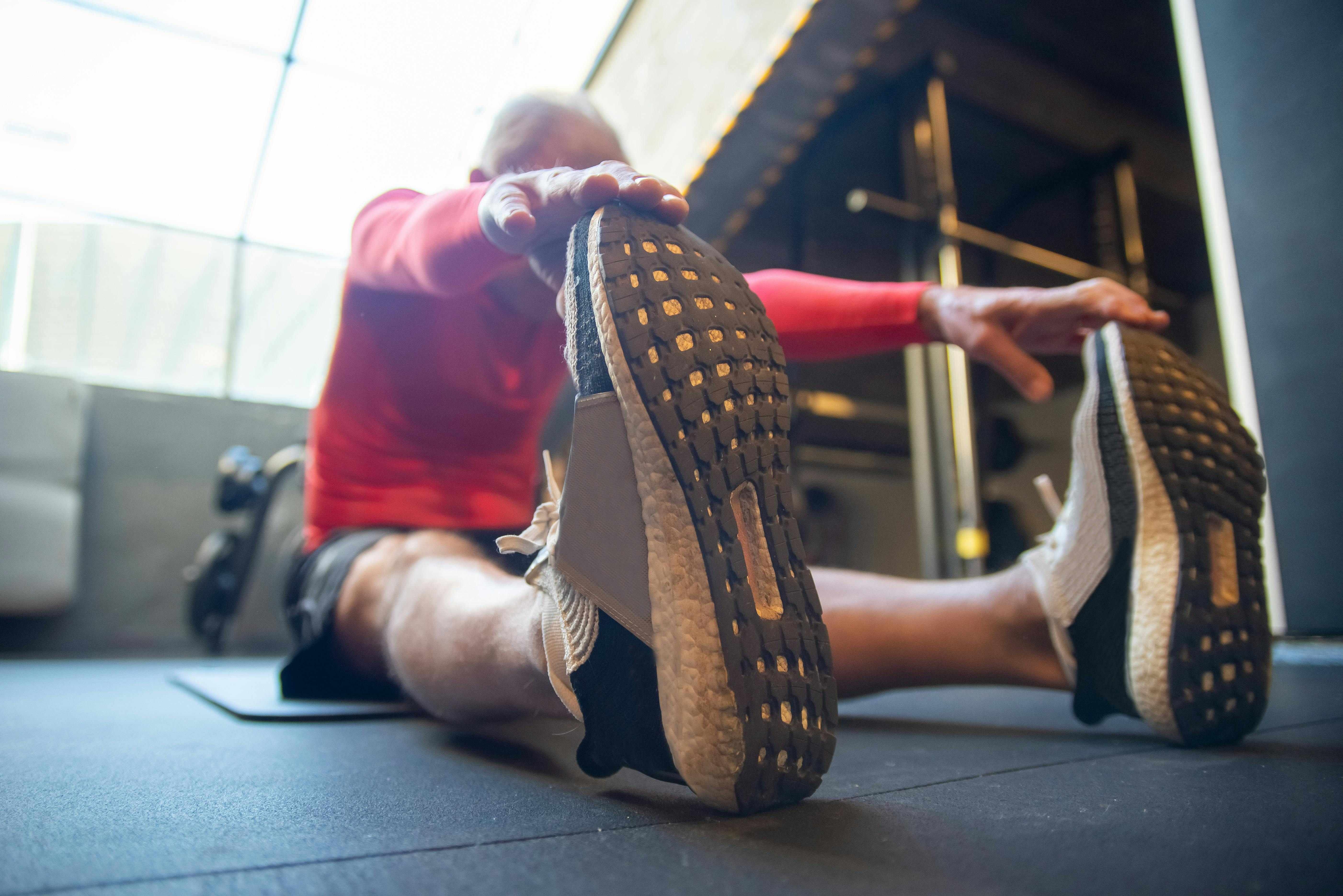 An older man stretching on a gym floor, touching his sneaker with his hands,demonstrating flexibility and proper form in a well-lit, community-focused gym environment.