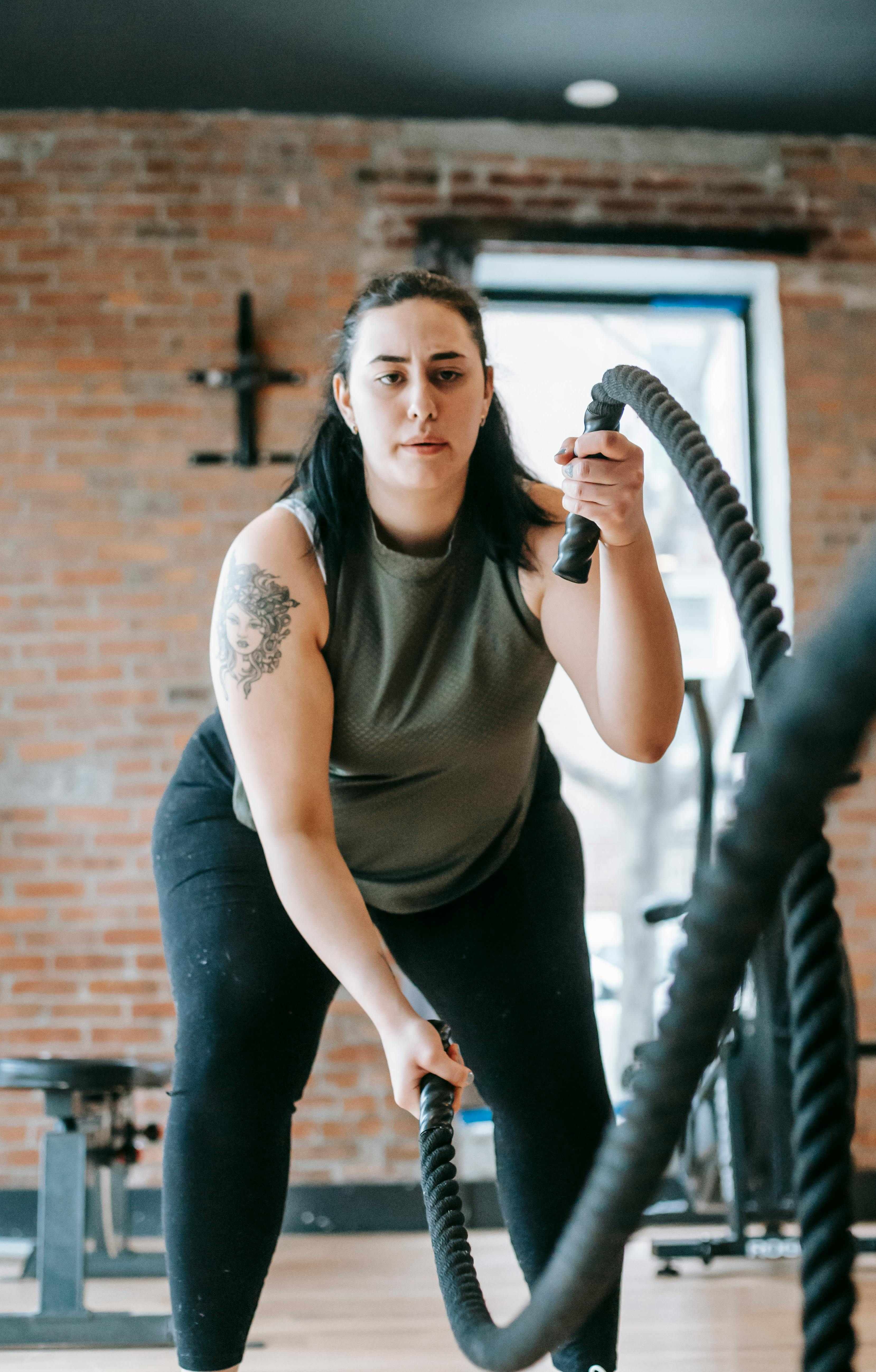 A woman with a tattoo, wearing a green tank top and black leggings, exercises with battle ropes in a gym. The gym has exposed brick walls and fitness equipment in the background. She is focused and determined, representing personalized training at Mojo Fitness Littleton.