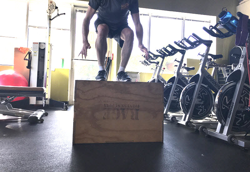 Person performing a box jump exercise in a gym setting, surrounded by fitness equipment like stationary bikes and weights. The image emphasizes strength and conditioning training, highlighting explosive power and lower body strength.