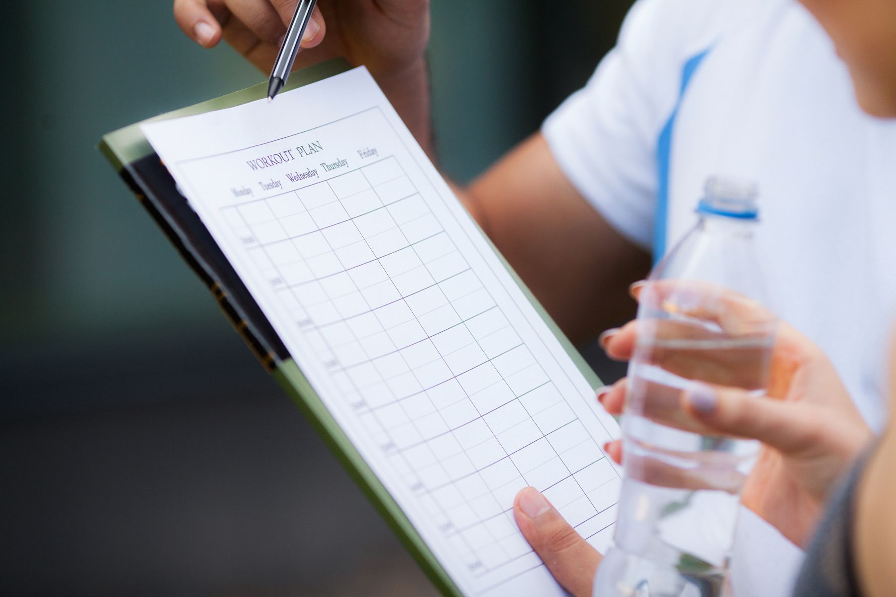 Person holding a workout plan on a clipboard and a water bottle, representing planning and consistency in fitness routines.