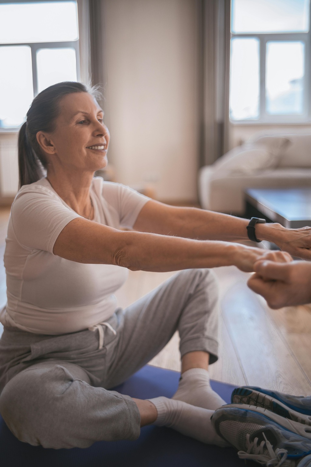 Senior woman stretching at Mojo Fitness Littleton, smiling while stretching her arms forward, wearing a white t-shirt and gray sweatpants.